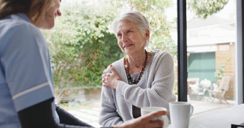 An older woman managing chronic pain holds her shoulder while talking with an in-home caregiver.
