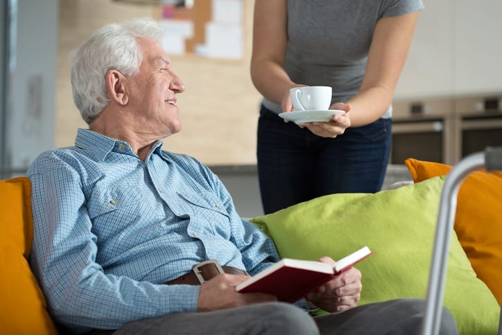 An older man reads a book and is handed a cup of tea by a caregiver who is helping him with recovery after surgery.
