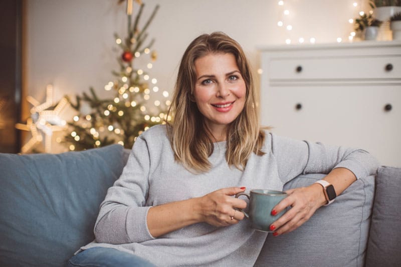A woman who knows it’s important to care for yourself relaxes with a mug of cocoa in a room that is warmly decorated for the holidays.
